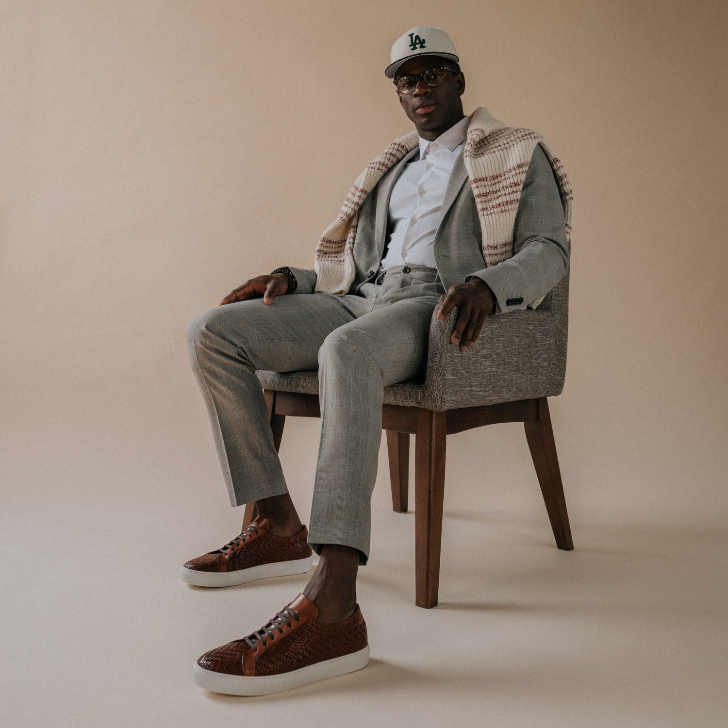 A man sits in a grey chair, wearing a grey suit, brown shoes, a white shirt, a beige and white checkered scarf, and a white cap with LA on it.