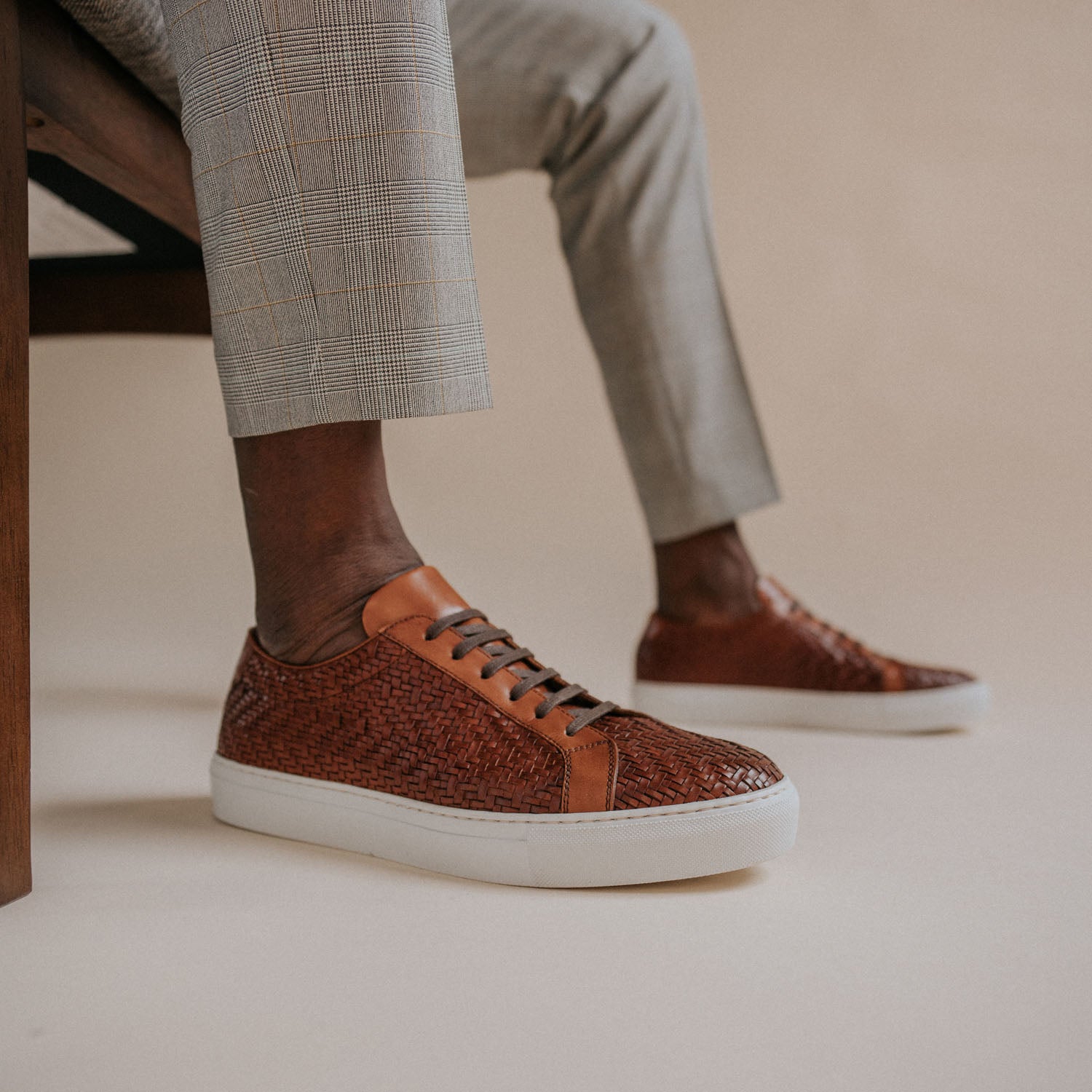 A person wearing plaid pants sits on a chair, showcasing brown woven leather sneakers with white soles against a plain background.