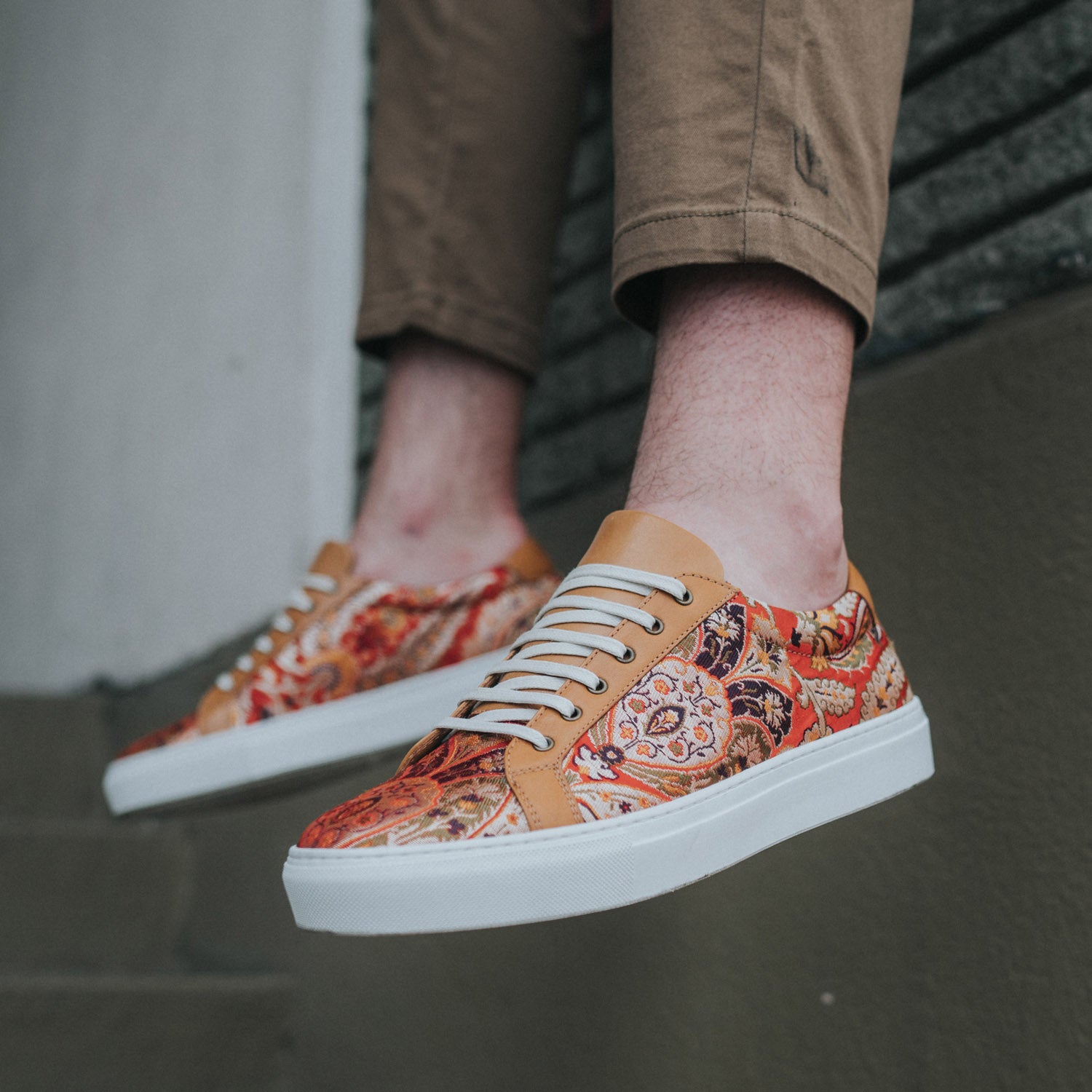 A person wearing brown patterned sneakers with white laces and white soles is seen standing on a staircase. They are also wearing brown pants.