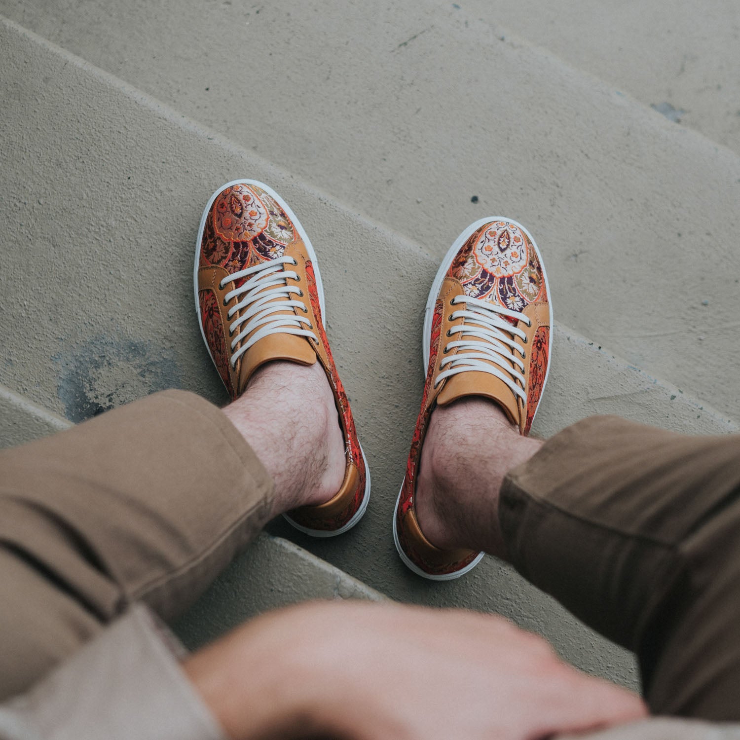 A person wearing colorful, patterned sneakers and light brown pants is seated on gray concrete steps. Only the lower legs and feet are visible.