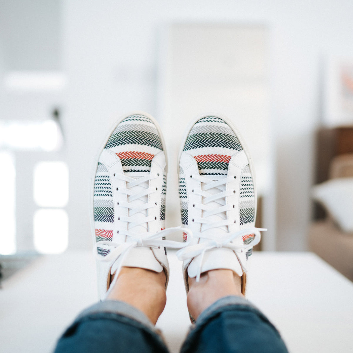 Person wearing white sneakers with multicolored patterns while resting feet on a surface, shown from a top-down perspective.