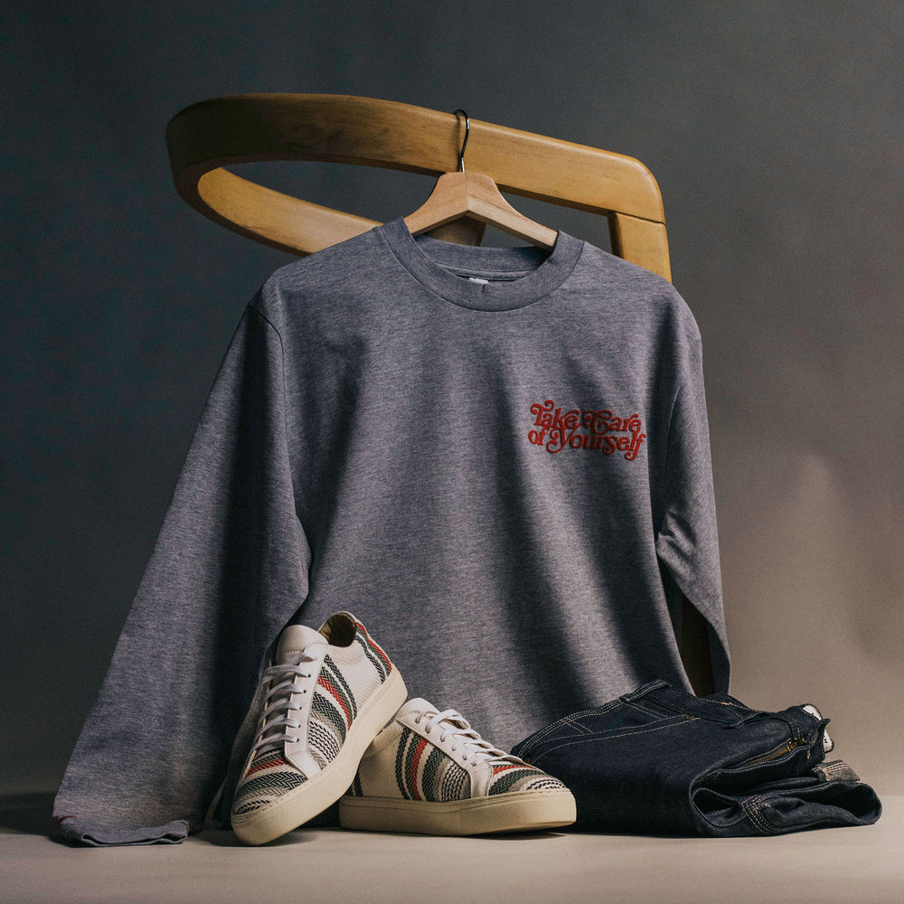 A gray long-sleeved shirt with text, striped sneakers, and dark jeans are displayed on a wooden chair against a plain background.