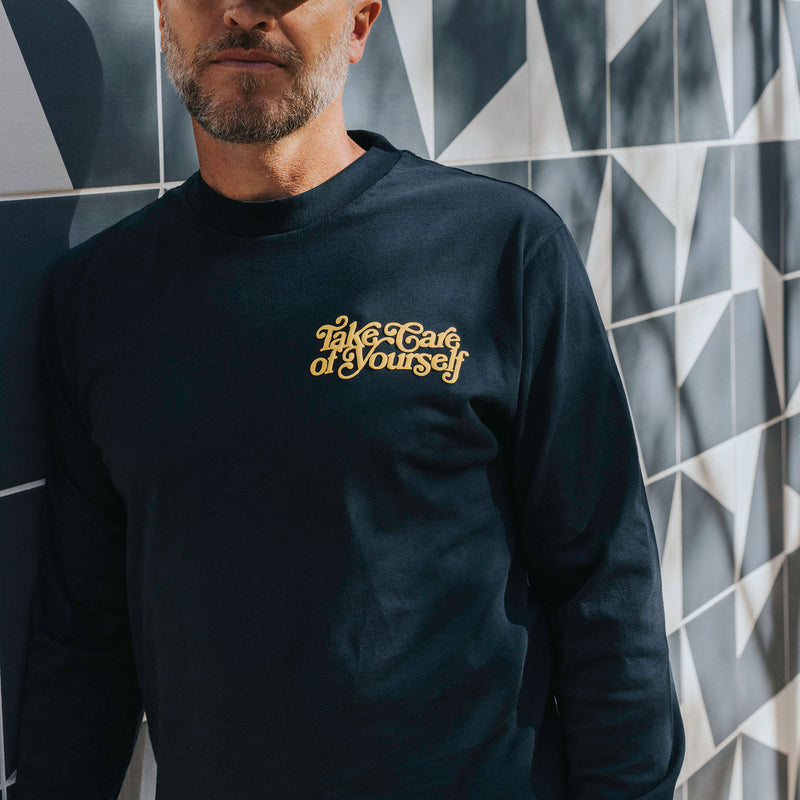 A man standing against a geometric patterned wall wears a black long-sleeve shirt with the phrase Take Care of Yourself printed in yellow on the chest.