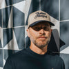 A man with a beard wears a beige cap with Salud Mental text, standing against a geometric patterned wall.