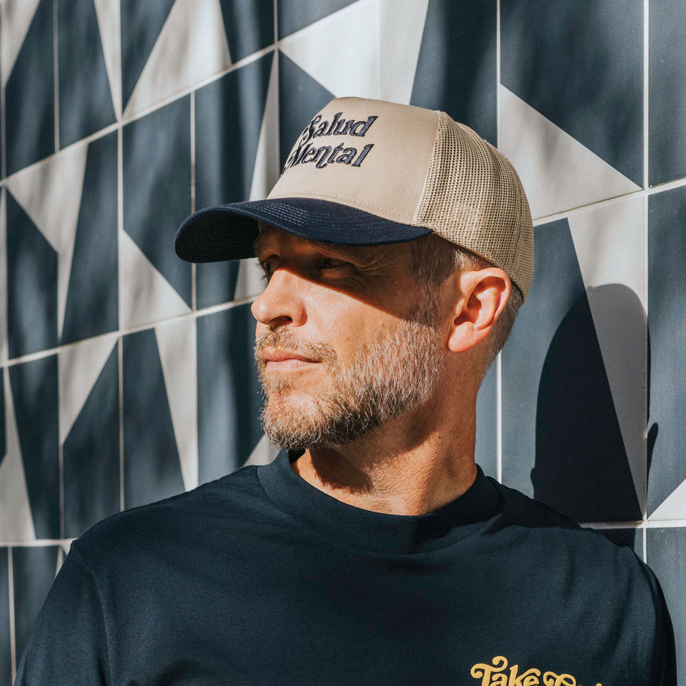 A man with a beard and mustache wearing a beige and black trucker hat and a navy blue shirt stands in front of a geometric black and white tiled wall.