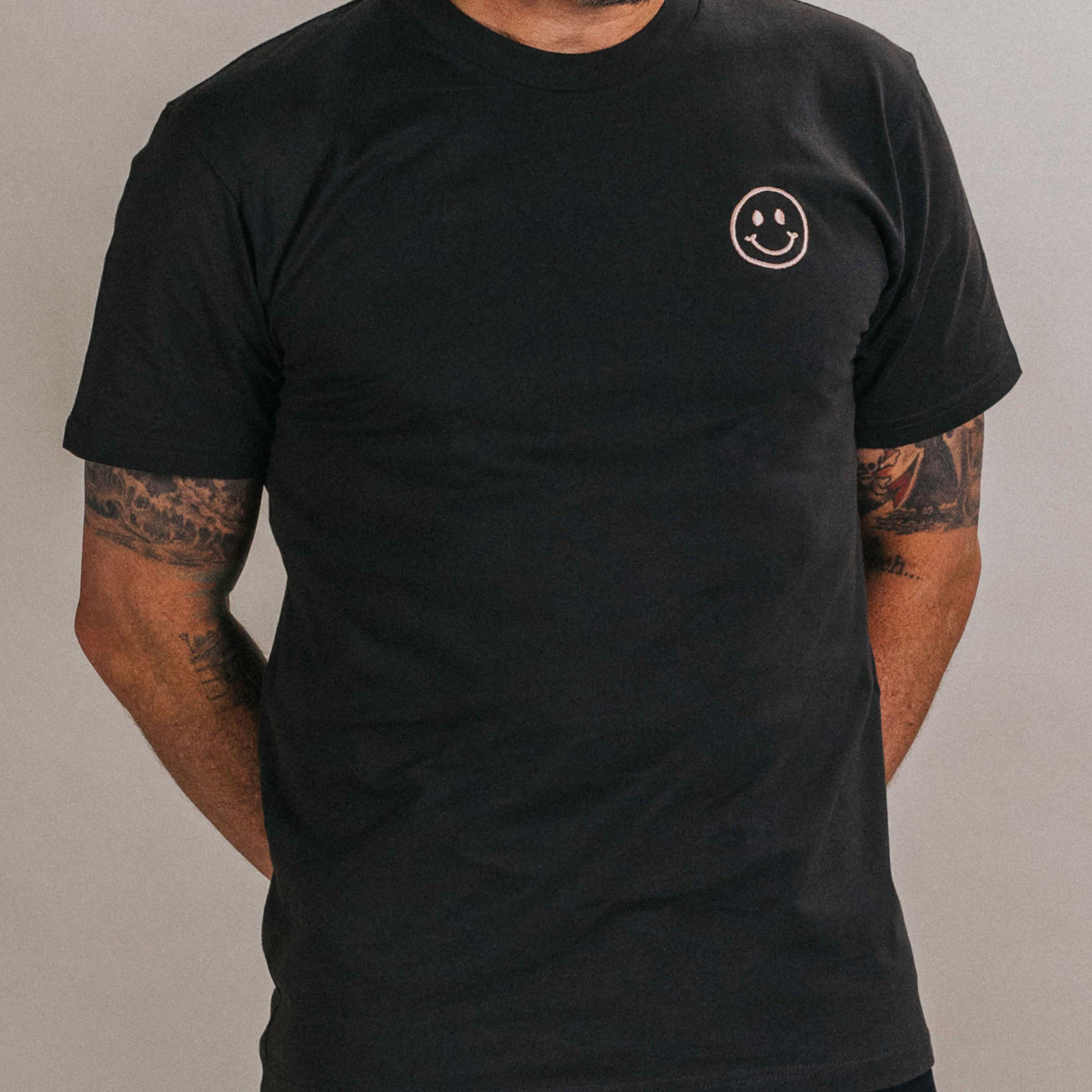 Person wearing a black T-shirt with a small smiley face logo on the upper left side, standing against a plain background.