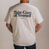 Back view of a person wearing a white T-shirt with the text Take Care of Yourself and TAFT written on the back. The person has visible arm tattoos and is standing against a plain background.