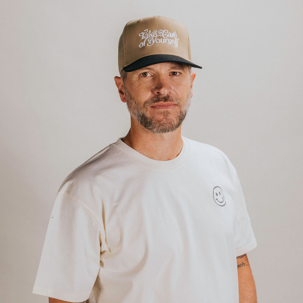 A man with a short beard and mustache, wearing a beige cap with text and a white t-shirt with a small smiley face emblem, stands against a plain background.