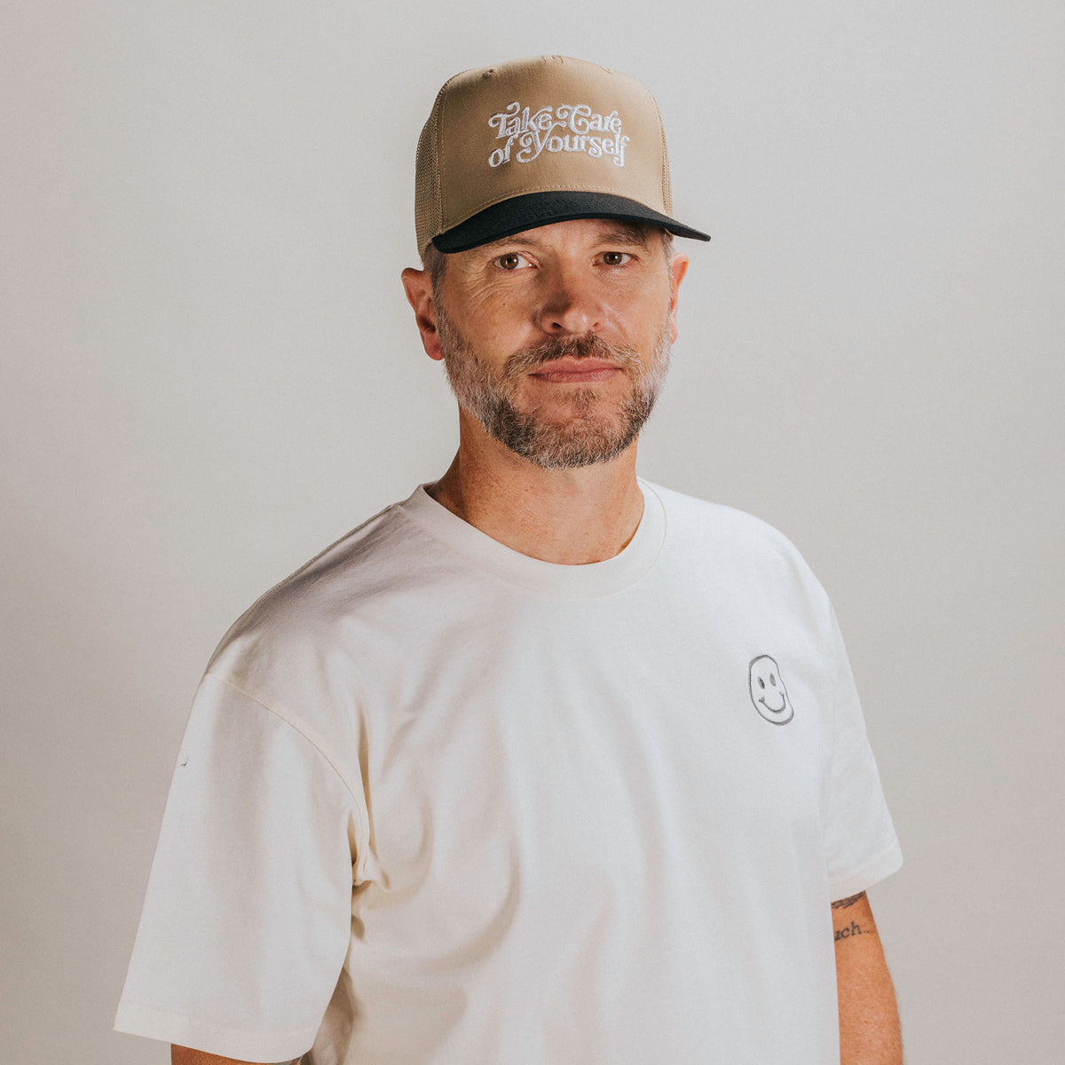 A man with a short beard and mustache, wearing a beige cap with text and a white t-shirt with a small smiley face emblem, stands against a plain background.