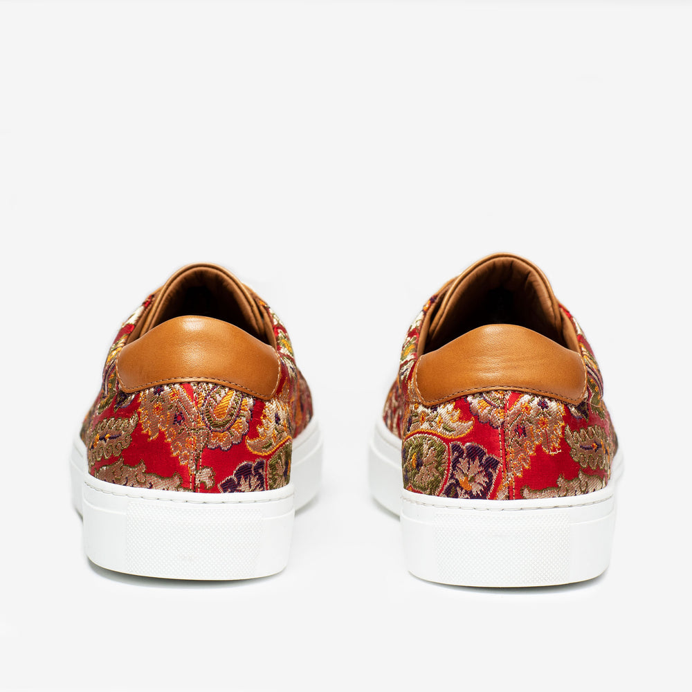 Rear view of a pair of patterned sneakers with a mix of red, gold, and purple fabric, and brown leather accents on the heel, complemented by white soles.