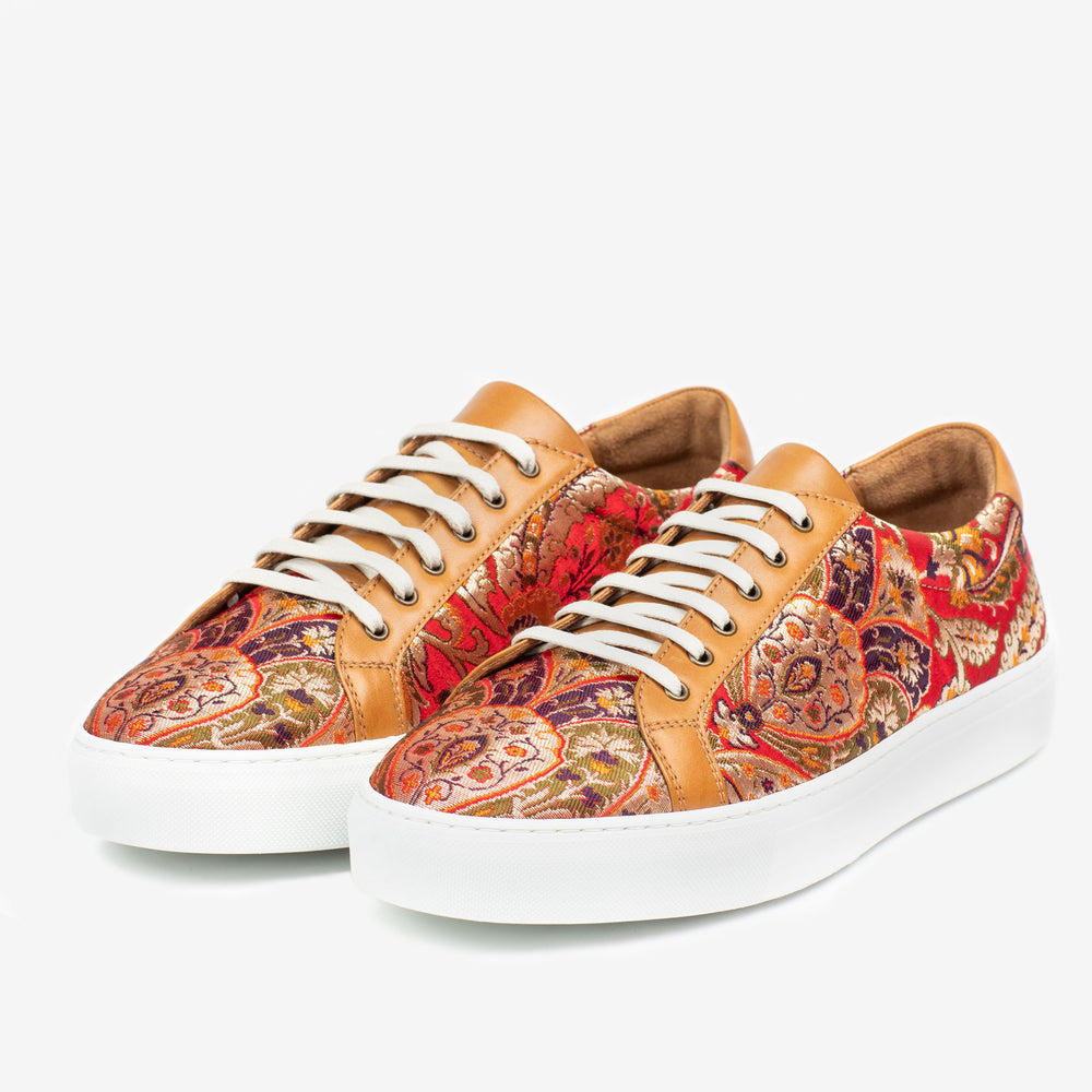 A pair of patterned sneakers featuring tan leather accents, white laces, and a white rubber sole. The intricate design includes red, orange, and purple hues.