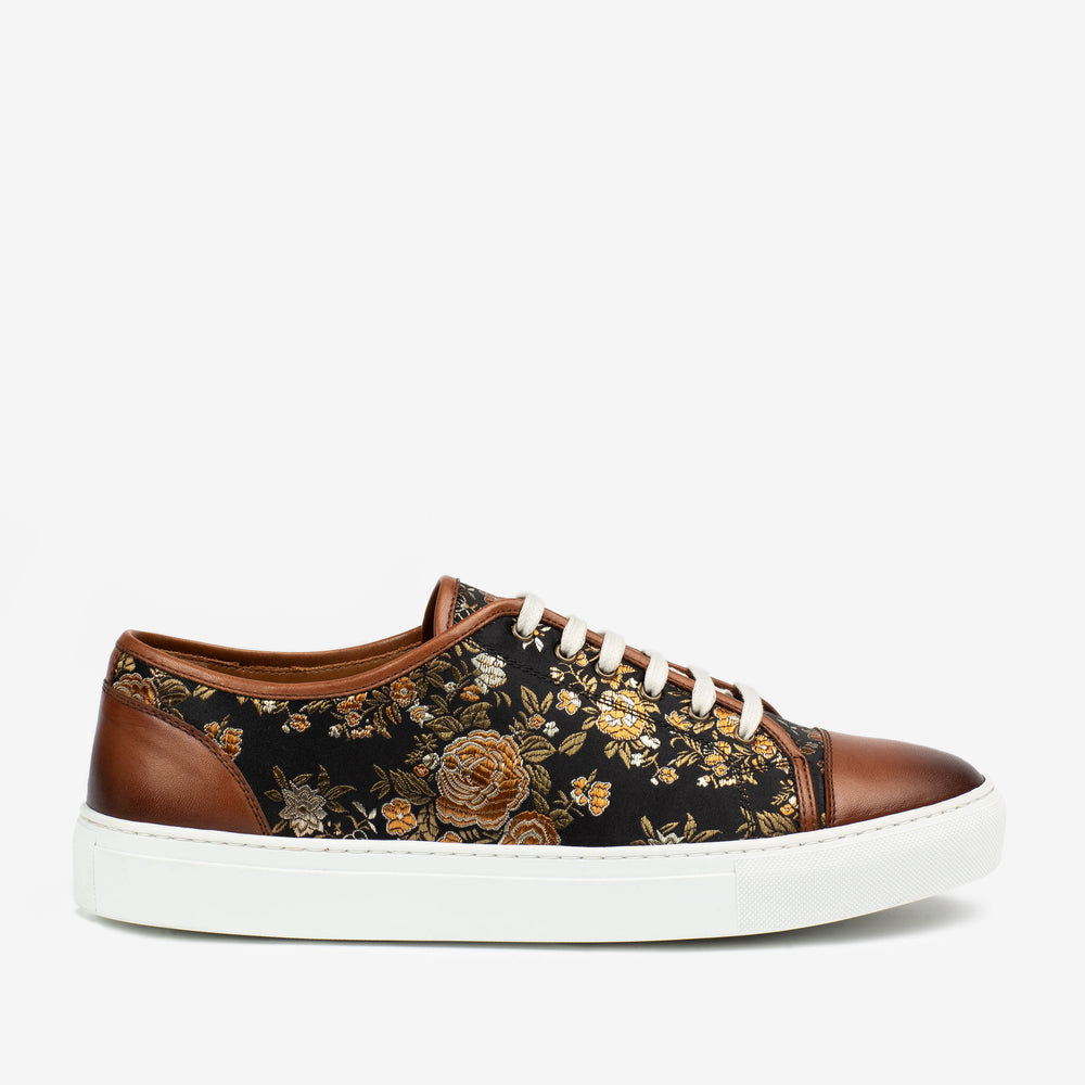 A low-top sneaker with a black fabric upper featuring a floral pattern and brown leather accents on the toe and heel, finished with white laces and a white rubber sole.