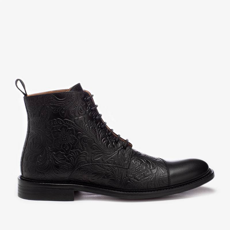 A black leather lace-up ankle boot with intricate embossed floral designs, featuring a pull tab at the back and a low heel.