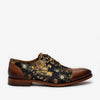 A black dress shoe with gold floral embroidery and brown leather accents on the toe and heel, isolated on a white background.