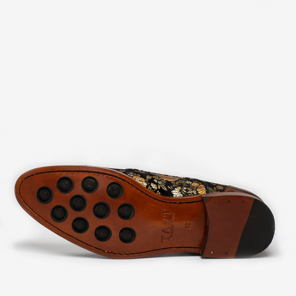 A leather dress shoe viewed from the bottom, showing a brown sole with eight round black grips and decorative floral embroidery on the upper part.