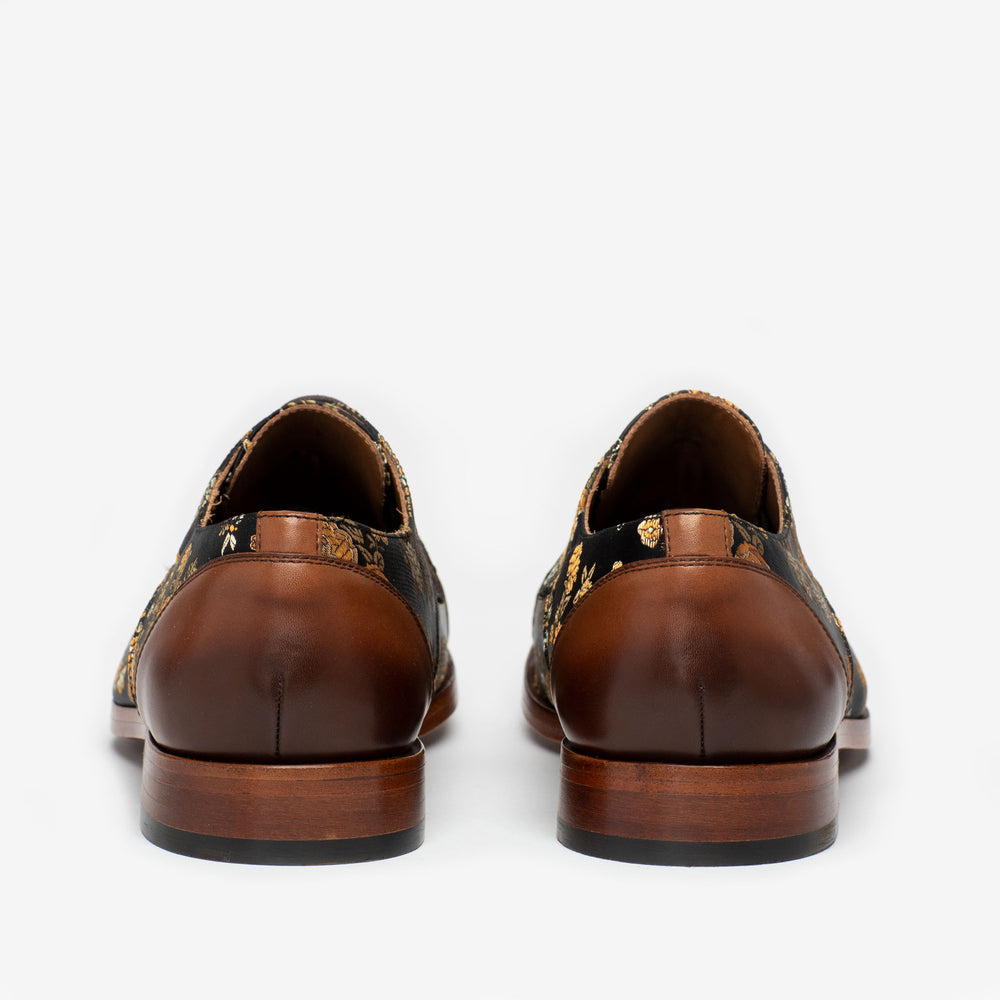 A pair of brown leather dress shoes with floral patterns on the back part, viewed from the rear.