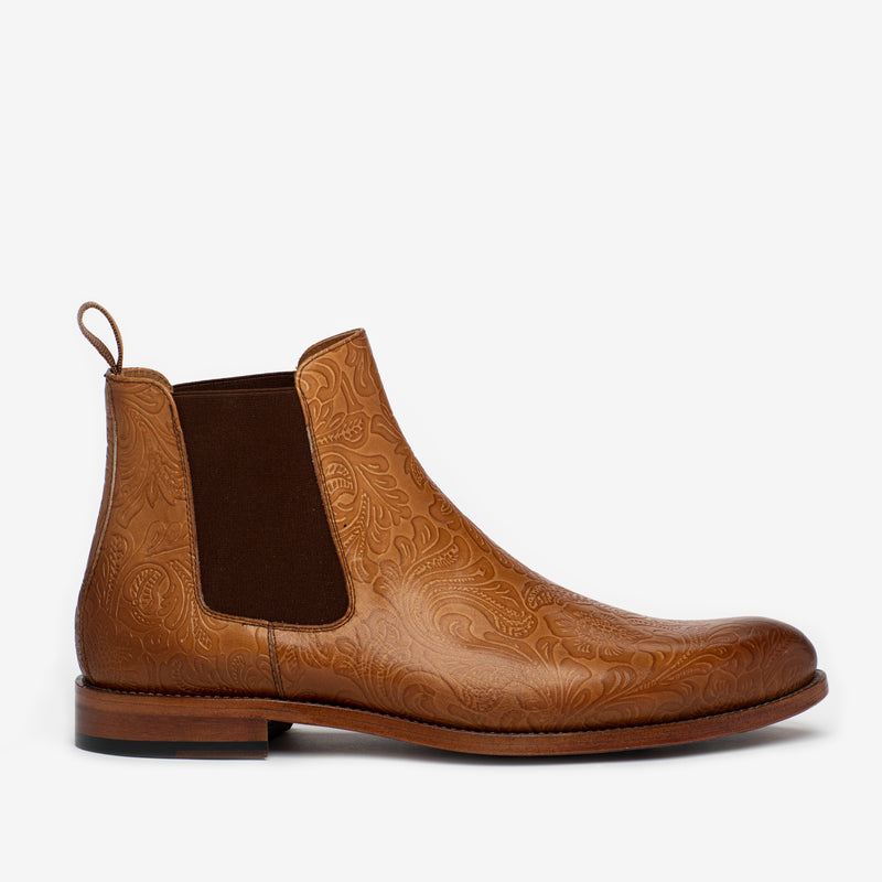 A single brown leather Chelsea boot with intricate embossed patterns, a pull tab, and elastic side panels, viewed from the side on a white background.