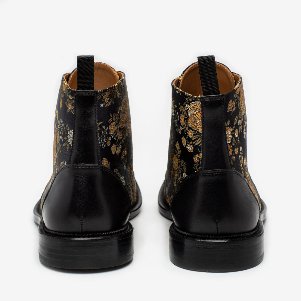 Rear view of a pair of black, leather and fabric ankle boots with a floral pattern and pull tabs on a white background.