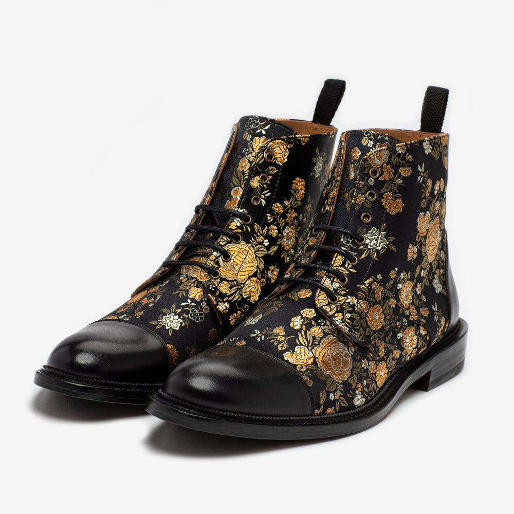 A pair of black leather ankle boots with a floral brocade pattern in gold and beige tones.