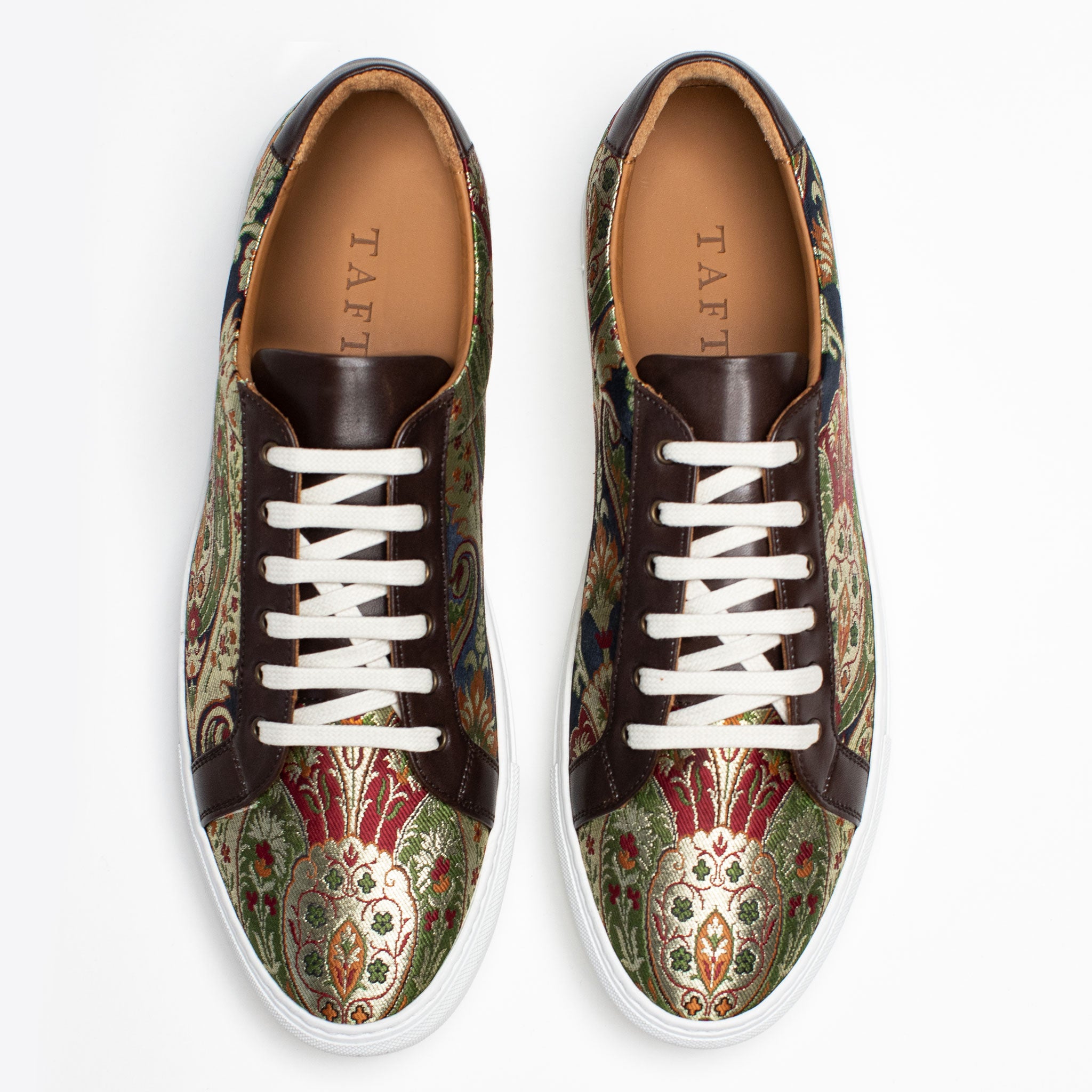 Top view of a pair of stylish sneakers featuring a paisley pattern in green, red, and gold colors, with brown leather accents and white soles and laces.