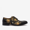 A single black dress shoe with a floral brocade pattern in gold and silver colors, featuring a black leather toe cap and low heel, viewed from the side against a white background.