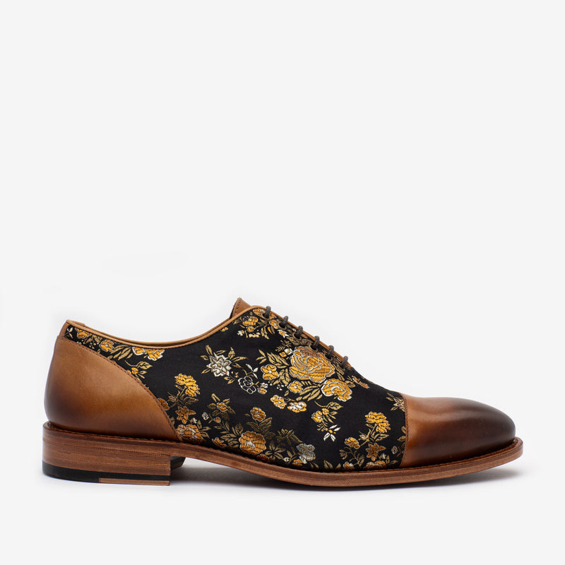 A brown leather dress shoe with a black fabric panel featuring gold and yellow floral designs.