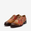 A pair of oxford-style shoes with brown leather toes and intricate red, gold, and beige paisley-patterned fabric on the rest.