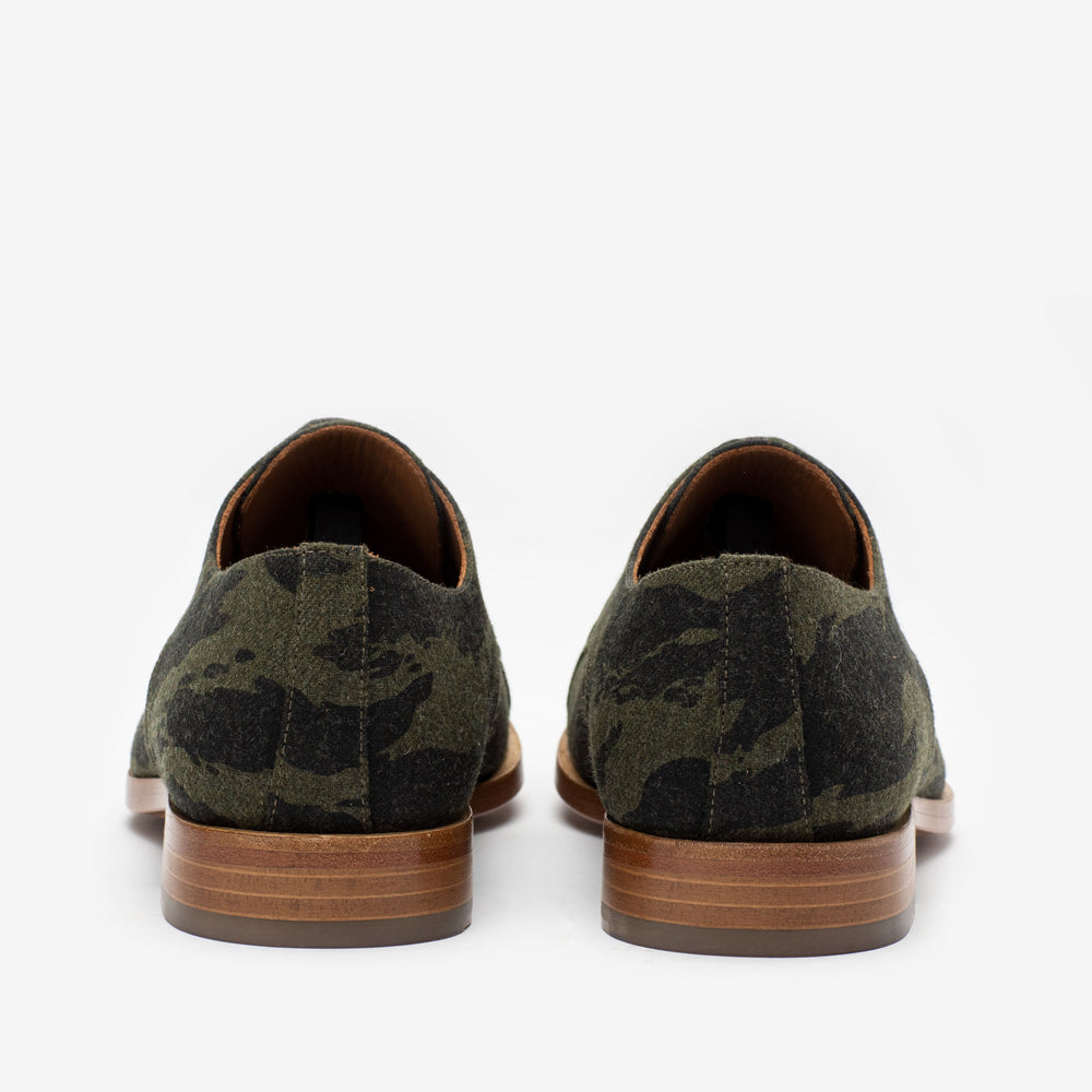 Rear view of a pair of olive green camo-patterned dress shoes with brown wooden soles, positioned against a white background.