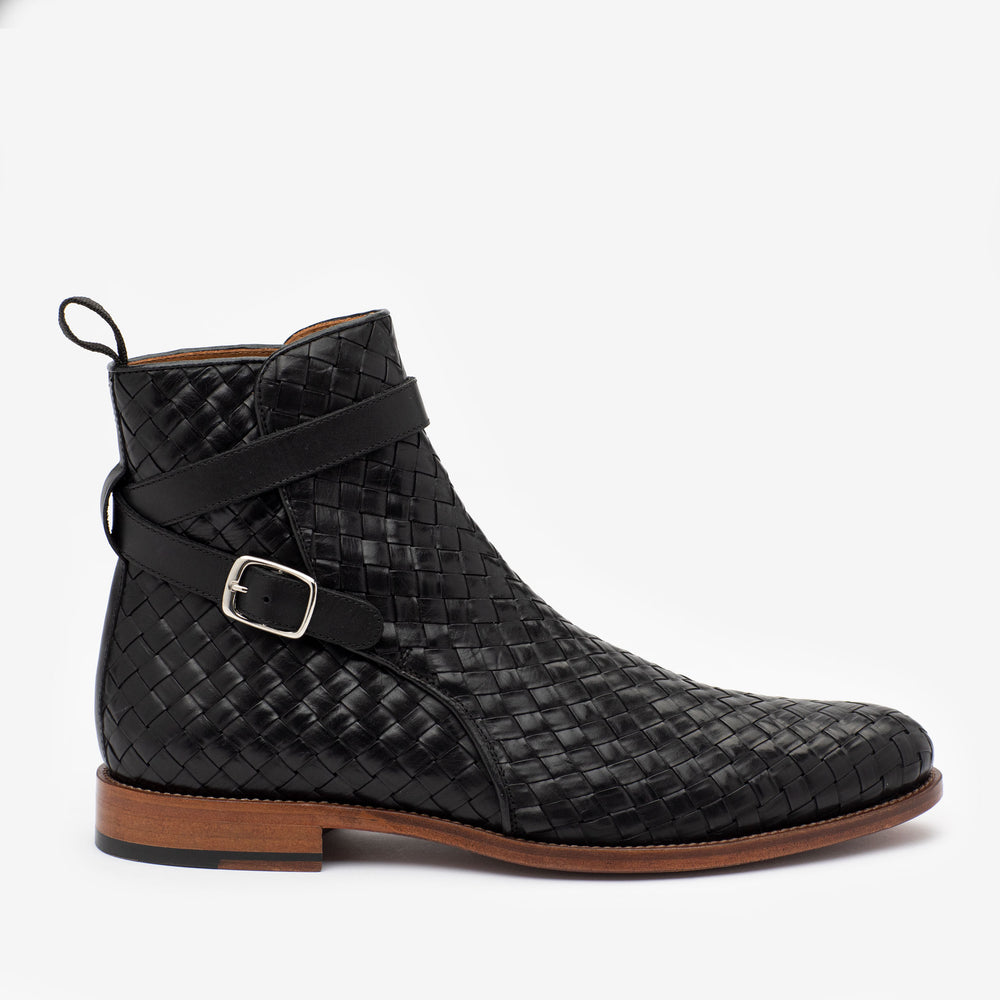 A black woven leather ankle boot with a buckle strap and wooden sole, viewed from the side against a white background.