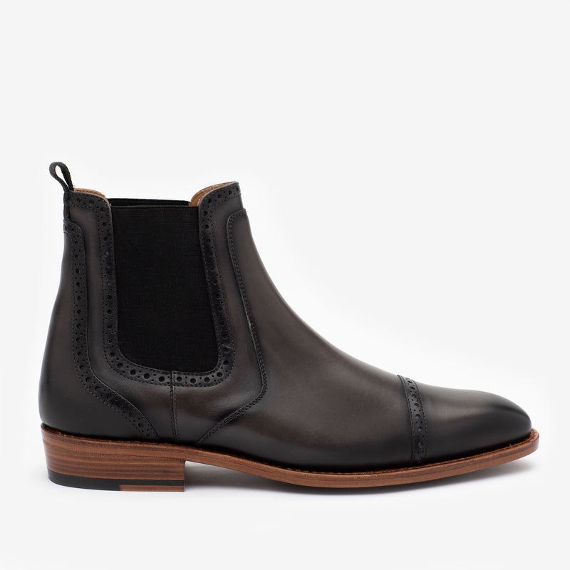 Black leather Chelsea boot with brogue detailing, elastic side panels, a pull tab at the back, and a wooden sole with a slight heel.