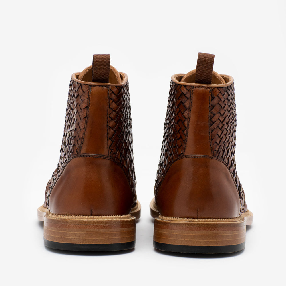 Rear view of a pair of brown leather boots with woven detailing, pull tabs, and wooden soles.