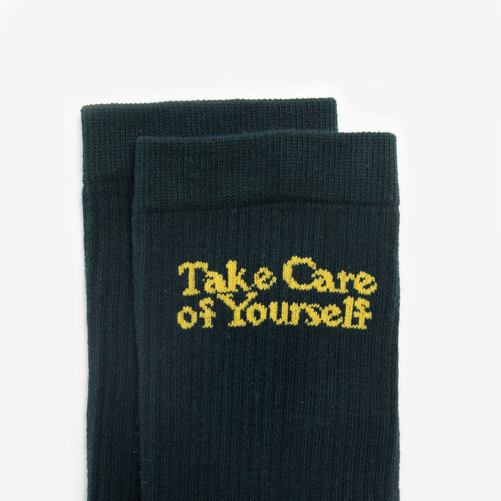 A pair of dark green socks with the text Take Care of Yourself in yellow embroidered on them.