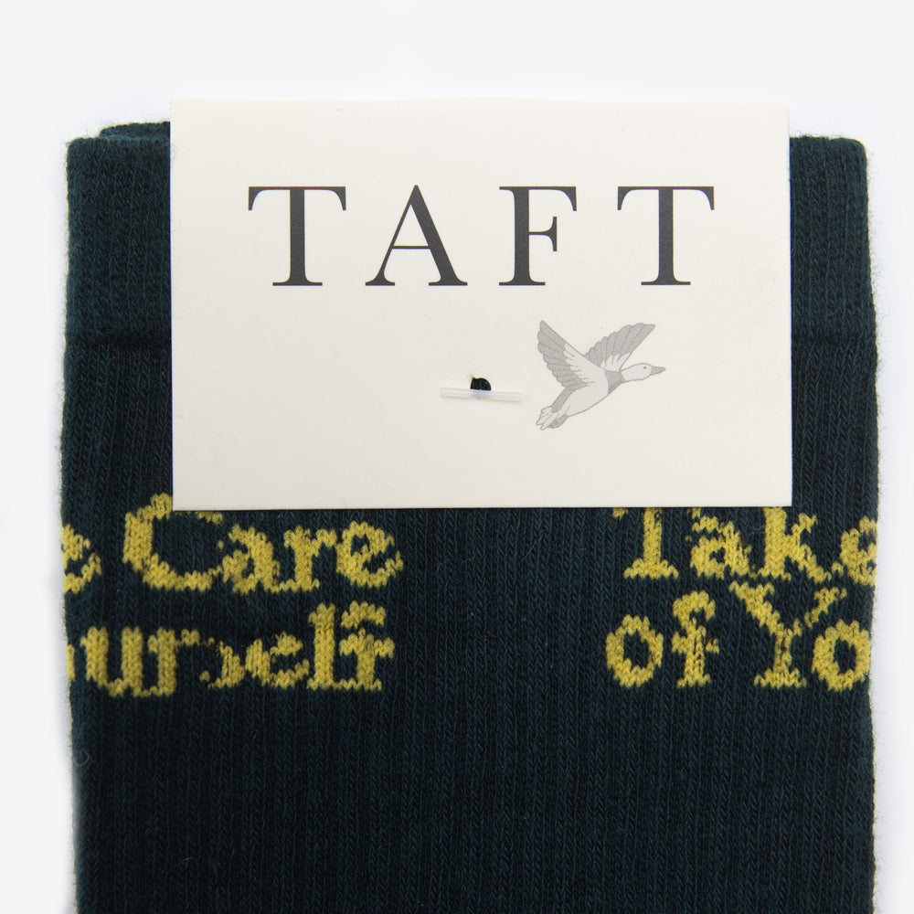 A dark green sock with the phrase Take Care of Yourself in yellow, partially covered by a white label with the brand name TAFT and an illustration of a bird.