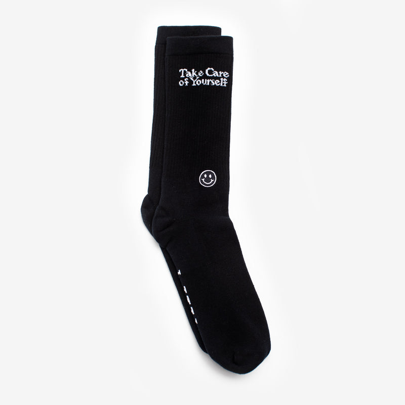 A pair of black socks with white text reading Take Care of Yourself near the top and a small white smiley face below it.