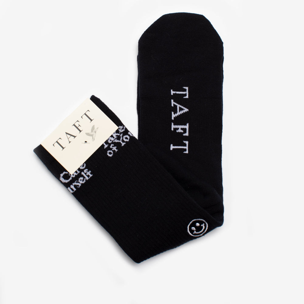 A pair of black socks with the brand name TAFT displayed on each sock. One sock is folded to show a card with the brand's logo.