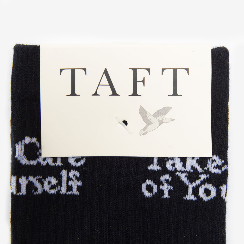 A pair of black socks with white text saying Take Care of Yourself, displayed with a TAFT brand tag featuring a flying bird graphic.