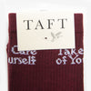 A pair of burgundy socks with white text partially visible under a packaging label that reads TAFT and features a bird image.