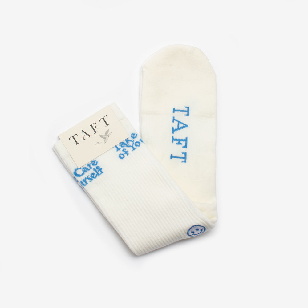 A pair of white socks with the brand name TAFT in blue. One sock is folded with a tag, and the other is laid flat showing the word TAFT and a smiley face at the cuff.