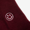 A close-up of a burgundy sock with a white smiley face embroidered on it. The fabric appears to be knit, and the design is simple and cheerful.