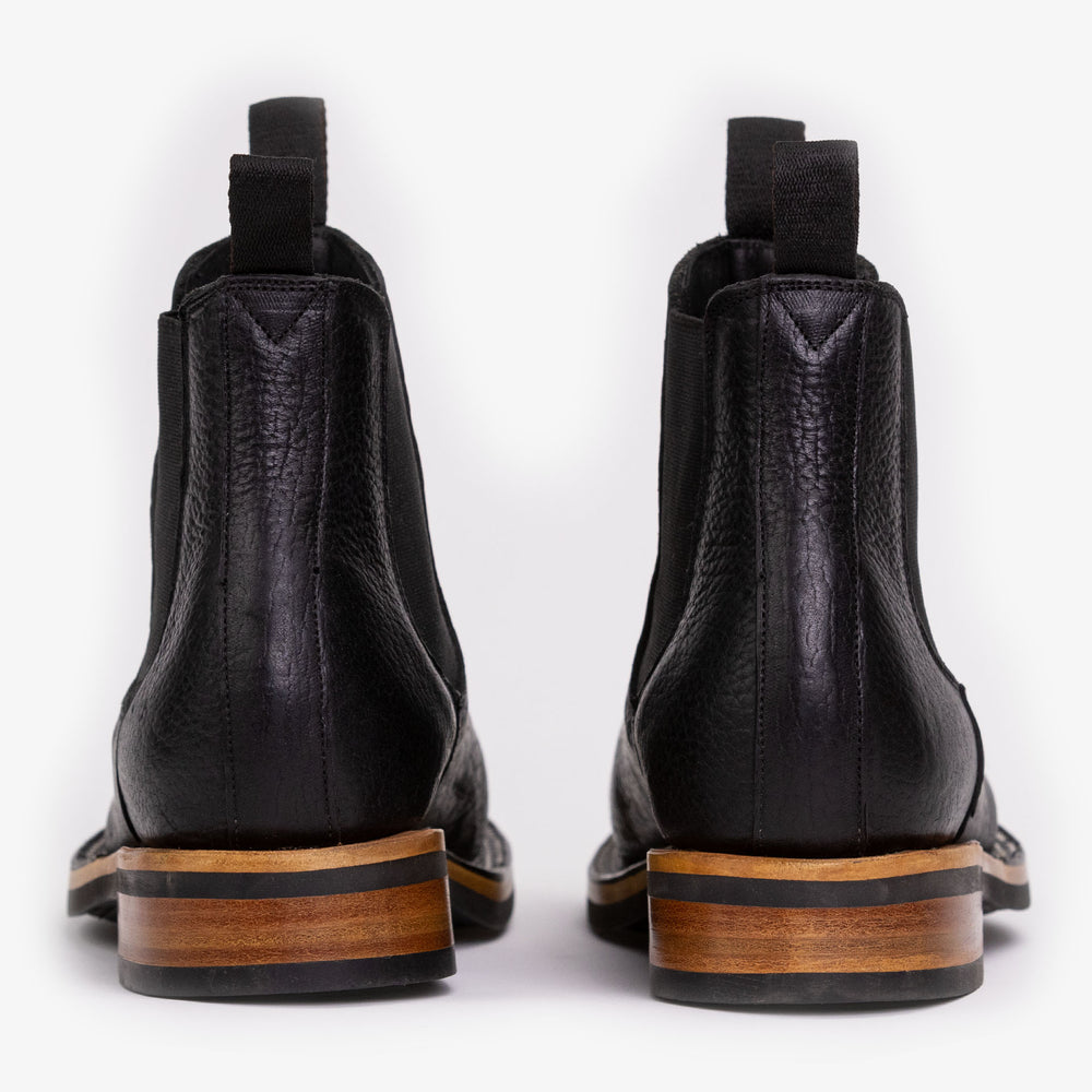 Rear view of a pair of black leather ankle boots with wooden soles, pull tabs, and elastic sides, placed side by side on a white background.