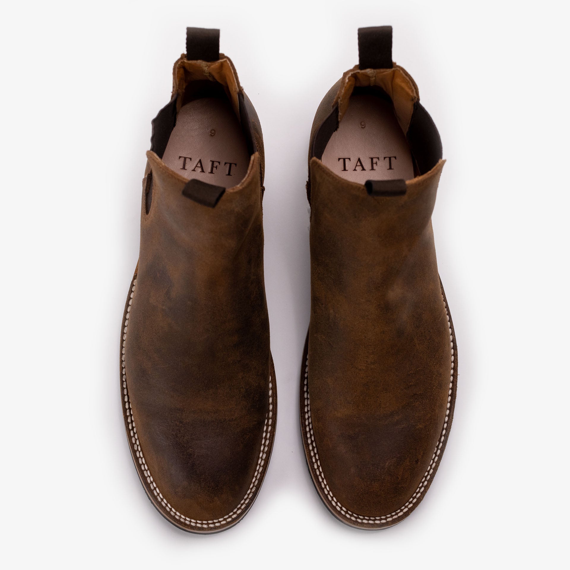 Top view of a pair of brown leather slip-on boots with a rounded toe and white stitching along the outsole. The brand name TAFT is visible on the insole.