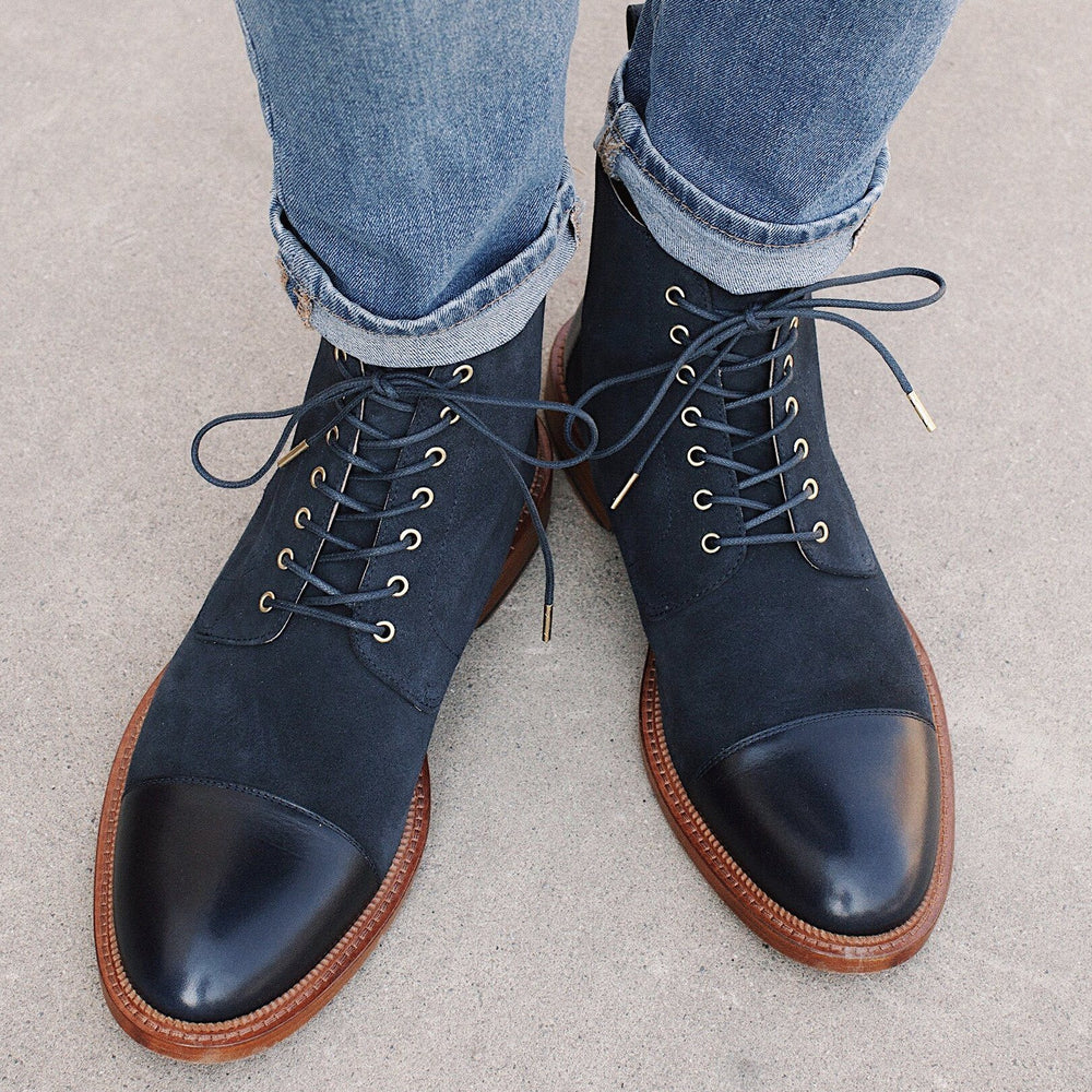 A person wears cuffed jeans and black, lace-up, leather cap-toe ankle boots on a concrete surface.