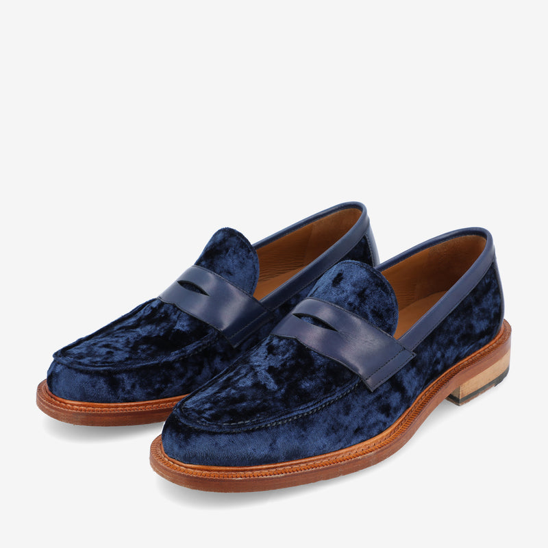 A pair of blue velvet loafers with a wooden sole and a leather strap across the front, set against a white background.