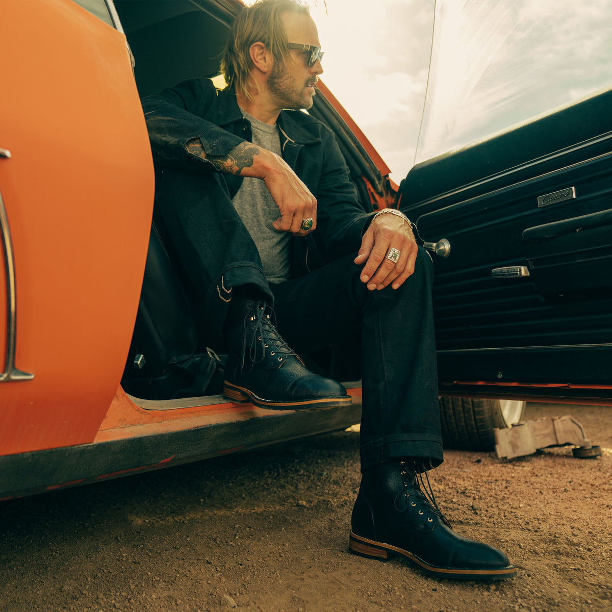 A person wearing sunglasses and sitting on the open door of an orange car, dressed in dark clothing and black boots, with a tattoo on the left arm.