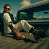 A person with sunglasses and a blue sweater sits casually in the bed of a truck, wearing white pants and brown boots.