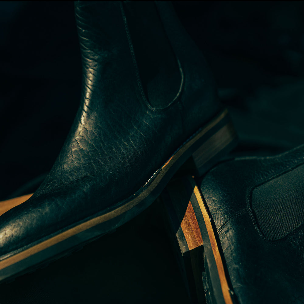 Close-up of two black leather boots with wooden soles, showcasing texture and detailing. The lighting is dark and moody.