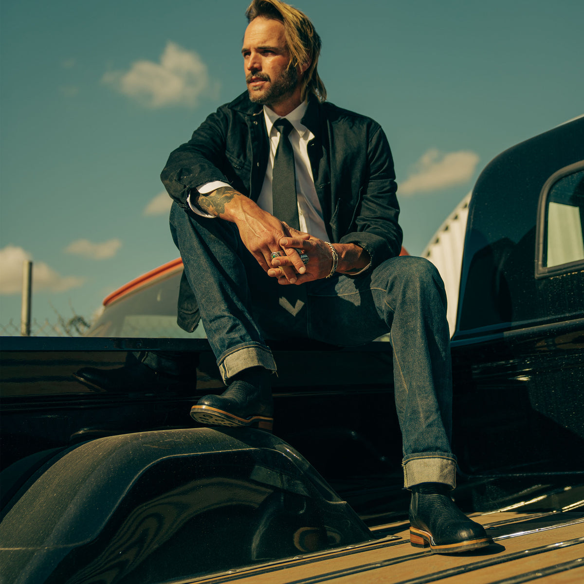 A man with long hair, wearing a black coat, white shirt, tie, and jeans, sits on the back of a pickup truck under a blue sky.