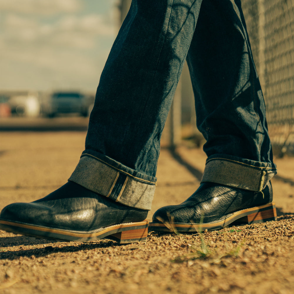 Close-up view of a person wearing dark blue cuffed jeans and black leather boots, standing on a dirt path with a chain-link fence in the background.