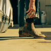 A person rolls up the cuff of their jeans, revealing brown leather boots and a ring on their hand, standing next to a vehicle on a sunlit concrete surface.