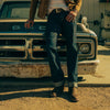 A person wearing jeans, boots, and a jacket leans against the front of an old, rusty truck parked on a dirt surface in an outdoor setting.